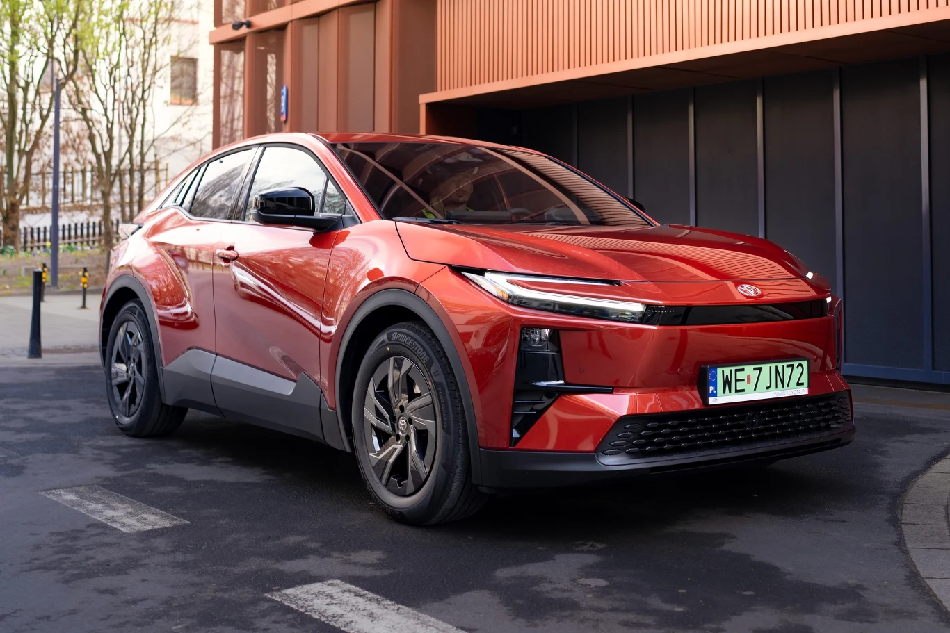 Toyota C-HR+ na jednym ładowaniu może przejechać do 607 km. Czerwony samochód elektryczny klasy SUV o dynamicznej sylwetce, nowoczesnym designie reflektorów i czarnych felgach, zaparkowany na ulicy miejskiej przy nowoczesnym budynku.