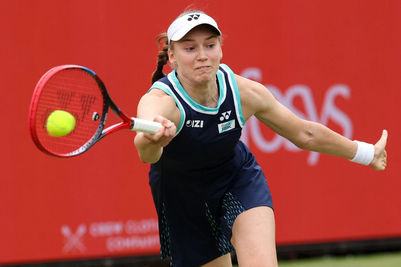 Jelena Rybakina, Queens 2026 Zawodniczka tenisowa w granatowym stroju i białej czapce uderza piłkę rakietą podczas meczu na tle czerwonego baneru reklamowego.
