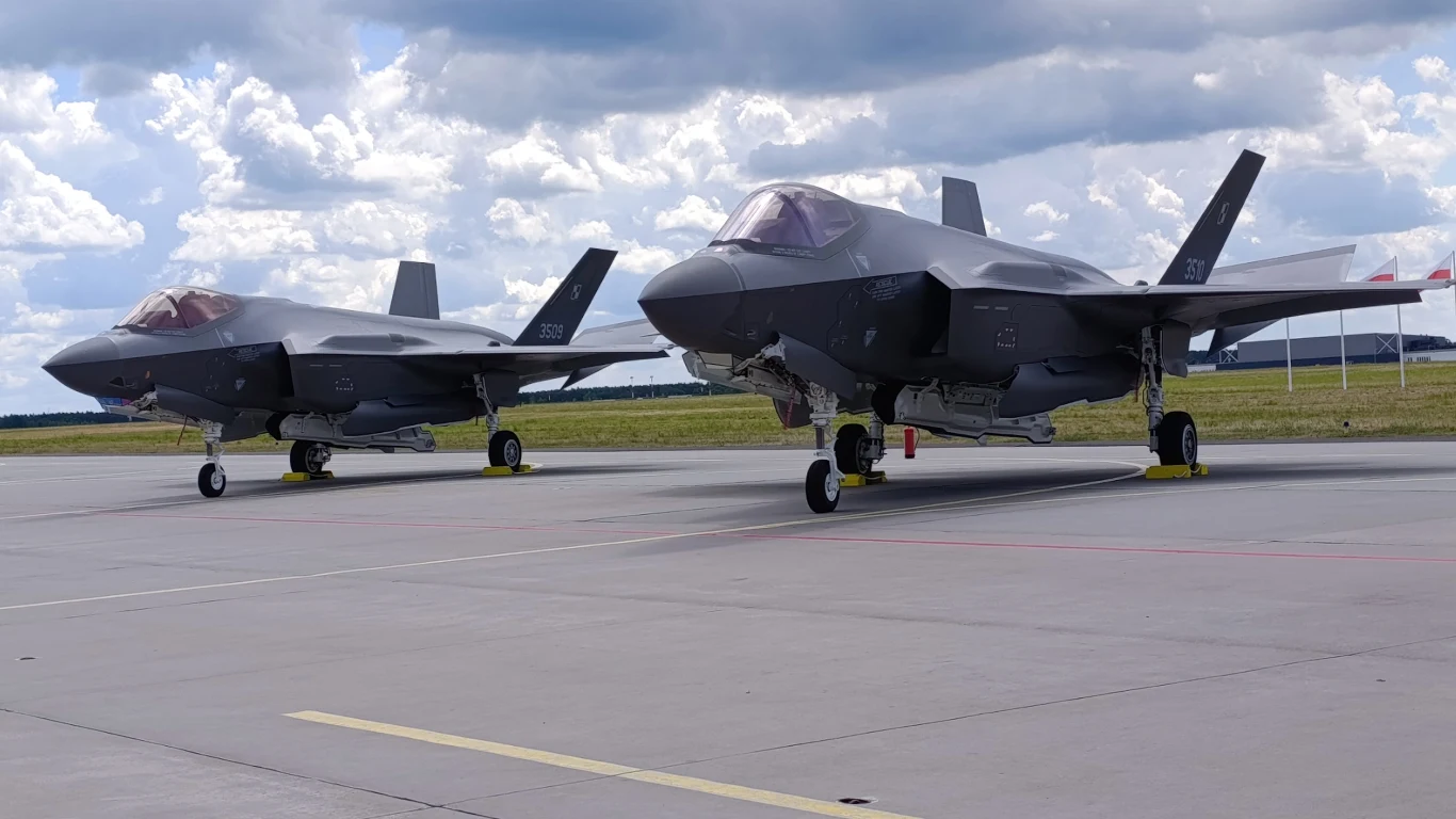 Dwa samoloty F-35 stojące obok siebie na płycie lotniska na tle pochmurnego nieba.