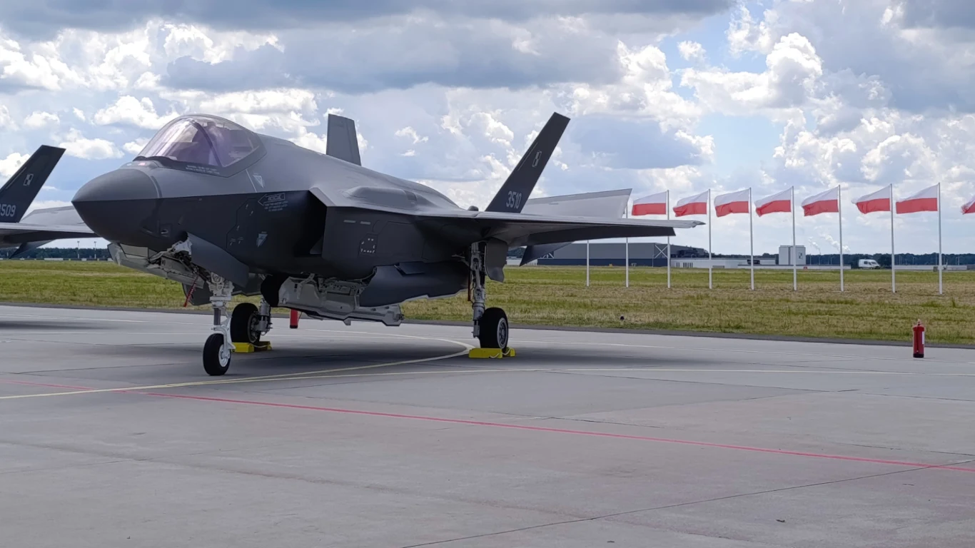 Myśliwiec F-35 ustawiony na lotniskowym pasie, w tle rząd powiewających polskich flag na masztach.