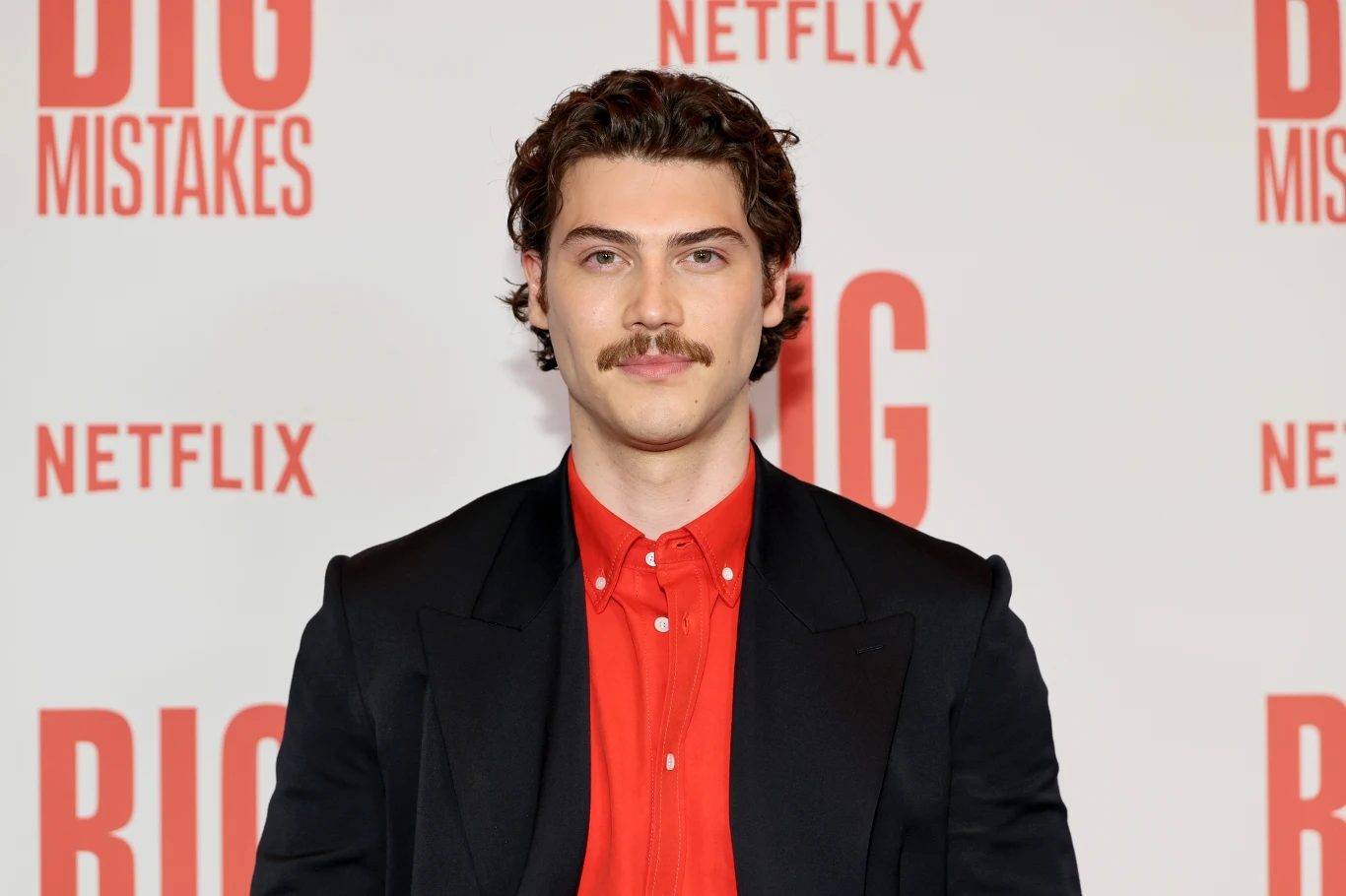 Mężczyzna z ciemnymi, kręconymi włosami i wąsem ma na sobie czarną marynarkę oraz czerwoną koszulę, stoi na tle z logo Netflix i napisem „BIG MISTAKES”.