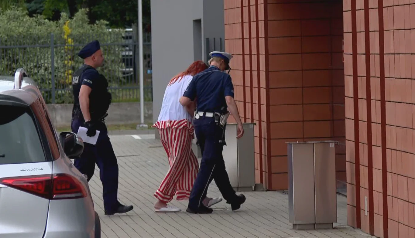 Dwóch policjantów prowadzi kobietę w biało-czerwonym stroju do budynku z czerwonej cegły.