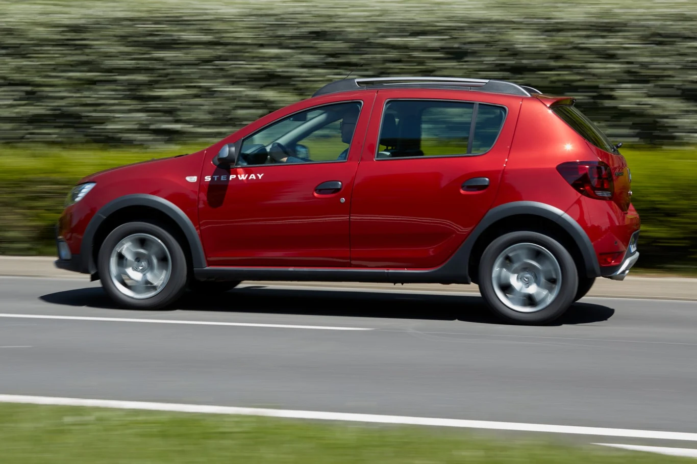 Czerwony samochód osobowy Dacia Sandero Stepway jedzie lewym pasem asfaltowej drogi, otoczony zielenią.