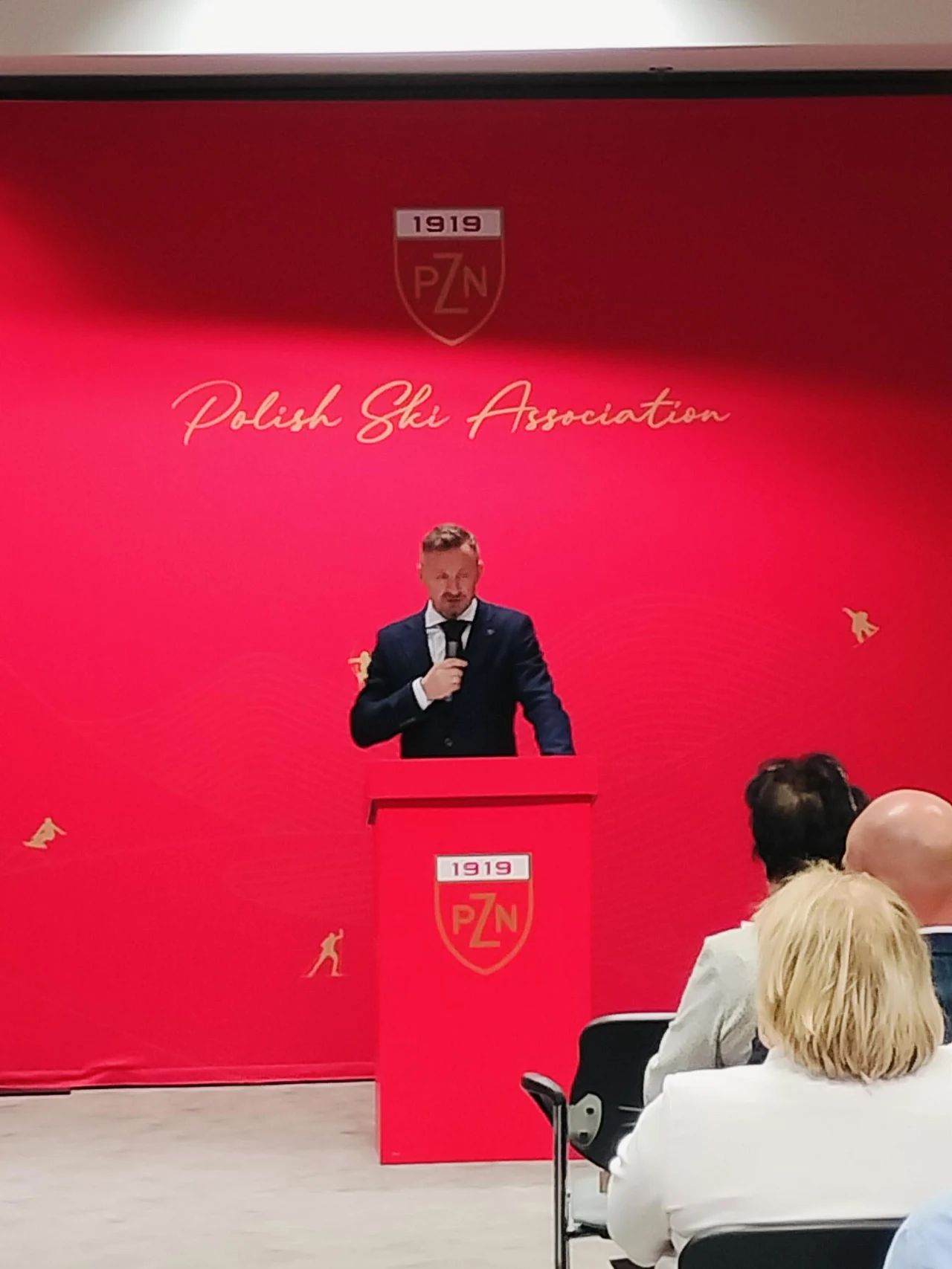 Mężczyzna w garniturze przemawia przy czerwonej mównicy z logo Polskiego Związku Narciarskiego, na tle czerwonej ścianki z napisem 'Polish Ski Association', przed słuchaczami siedzącymi w sali.