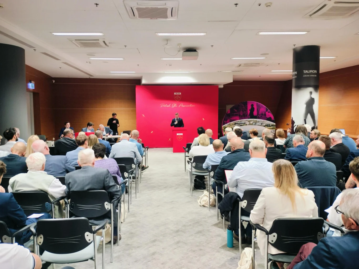 Prelegent przemawia ze sceny na tle różowej ściany z napisem, przed liczną publicznością siedzącą na krzesłach w sali konferencyjnej.