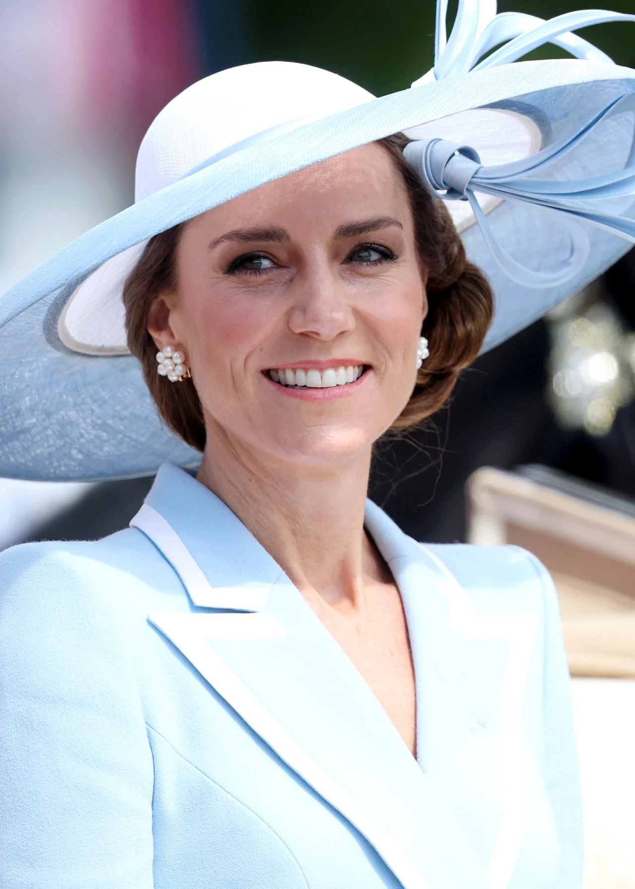 Księżna Kate Uśmiechnięta kobieta w eleganckim, jasnoniebieskim stroju i dużym kapeluszu z dekoracyjnym rondem, ozdobiona perłowymi kolczykami w kształcie kwiatów.