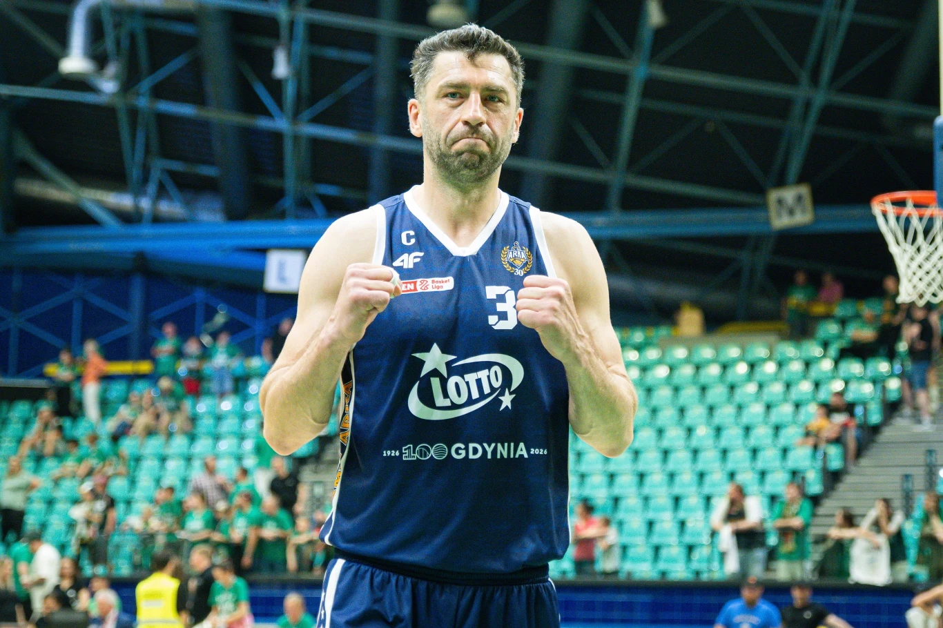 Koszykarz w granatowej koszulce z numerem 33, zaciskający pięści, stoi na tle trybun pełnych kibiców w hali sportowej.