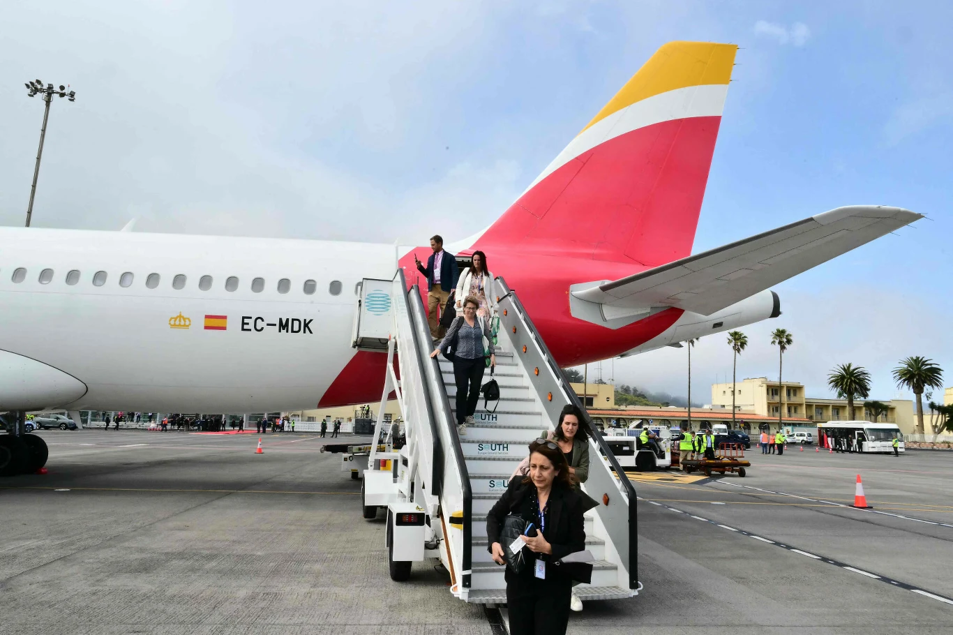 Pasażerowie schodzący po schodach z samolotu linii Iberia, który stoi na płycie lotniska w otoczeniu palm i zabudowań.