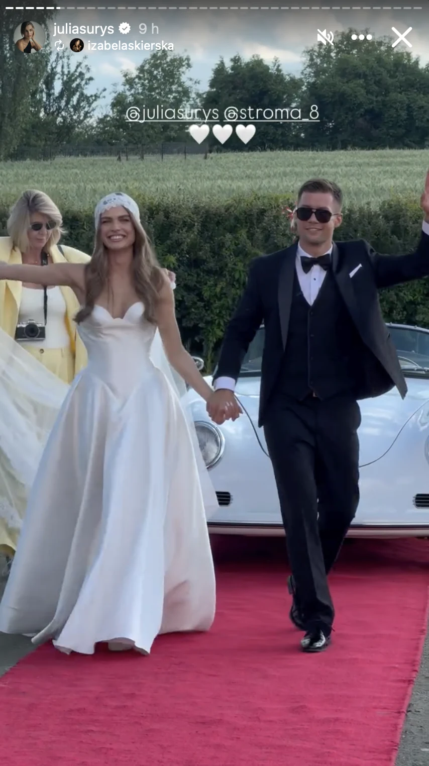 Para młoda w eleganckich strojach, uśmiechnięta i trzymająca się za ręce, przechodzi po czerwonym dywanie przy białym, klasycznym samochodzie.