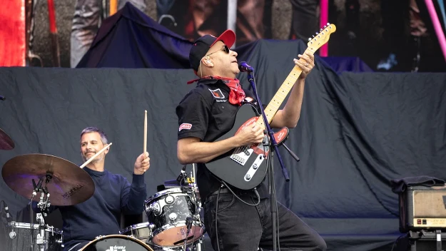 Gitarzysta w czapce i bandanie gra intensywnie na gitarze elektrycznej, obok niego perkusista uderza w bębny, obaj muzycy na tle sceny podczas koncertu.