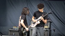 Dwóch młodych mężczyzn grających na gitarach elektrycznych na scenie koncertowej, w tle wzmacniacze i sprzęt muzyczny.
