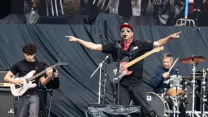 Trzech muzyków na scenie: gitarzysta z gitarą elektryczną po lewej, wokalista z gitarą i mikrofonem na środku w energicznej pozie oraz perkusista za zestawem perkusyjnym po prawej; dynamiczna atmosfera koncertu rockowego.