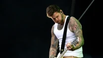 Wytatuowany mężczyzna w białej koszulce na ramiączkach gra na białej gitarze elektrycznej, pochylając głowę w skupieniu.