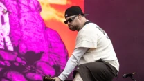 Mężczyzna w czapce z daszkiem, okularach przeciwsłonecznych i jasnej bluzie gra na gitarze elektrycznej na tle kolorowej, fioletowej grafiki przedstawiającej skały.
