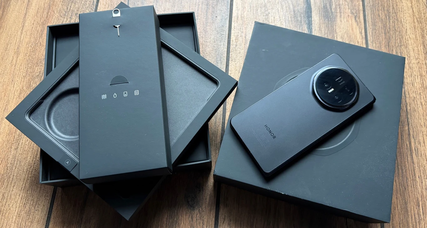 Czarny smartfon marki Honor z dużą, okrągłą wyspą aparatów, obok rozłożone czarne pudełka i akcesoria na drewnianej powierzchni.