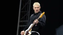 Mężczyzna z krótkimi, jasnymi włosami grający na białej gitarze basowej na scenie, ubrany w czarną koszulę i ciemne spodnie, z wyrazistą mimiką twarzy podczas występu.