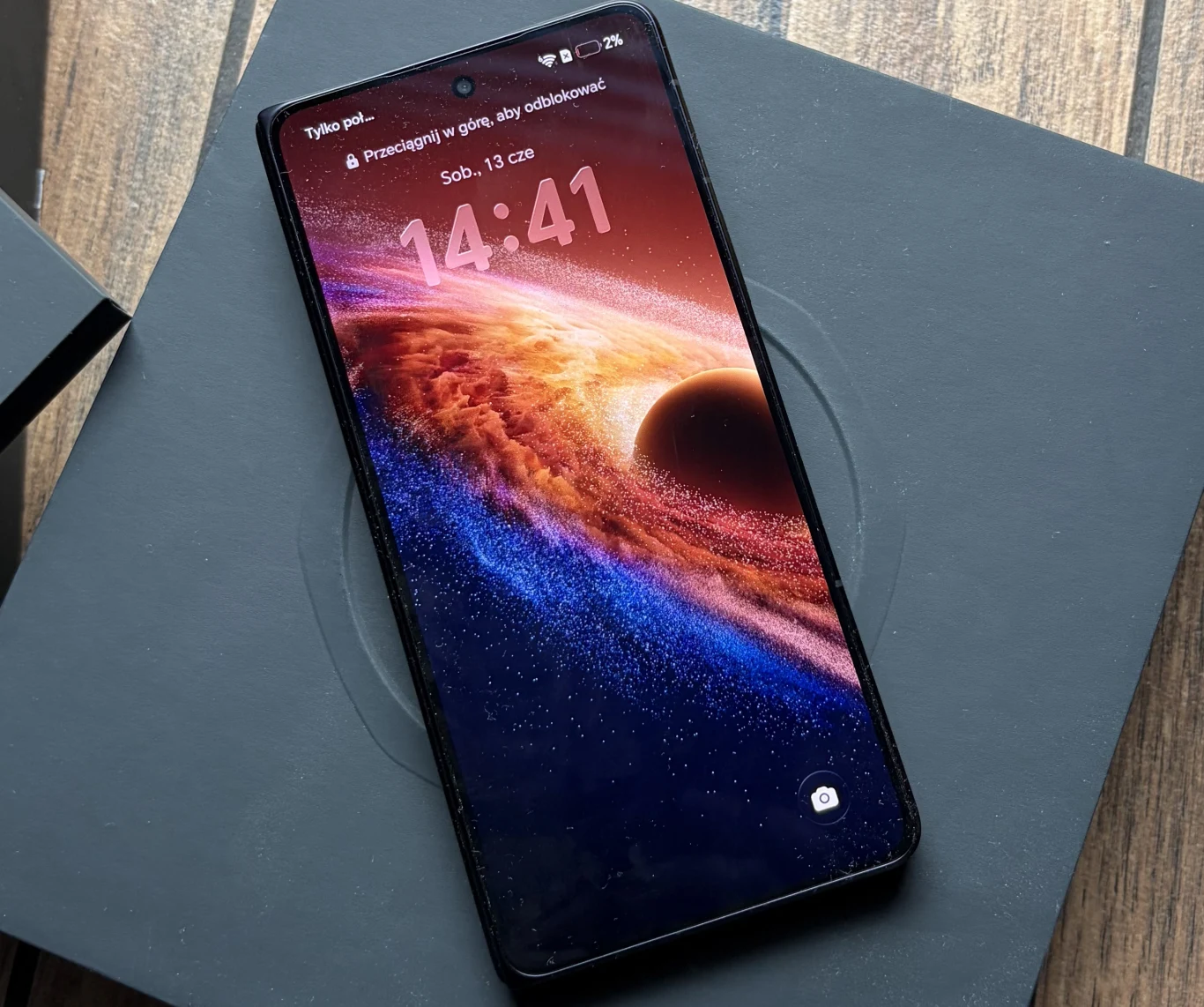 Czarny smartfon z wyświetlaczem pokazującym kosmiczną tapetę z planetą i pierścieniami, położony na ciemnym pudełku na tle drewnianej powierzchni.