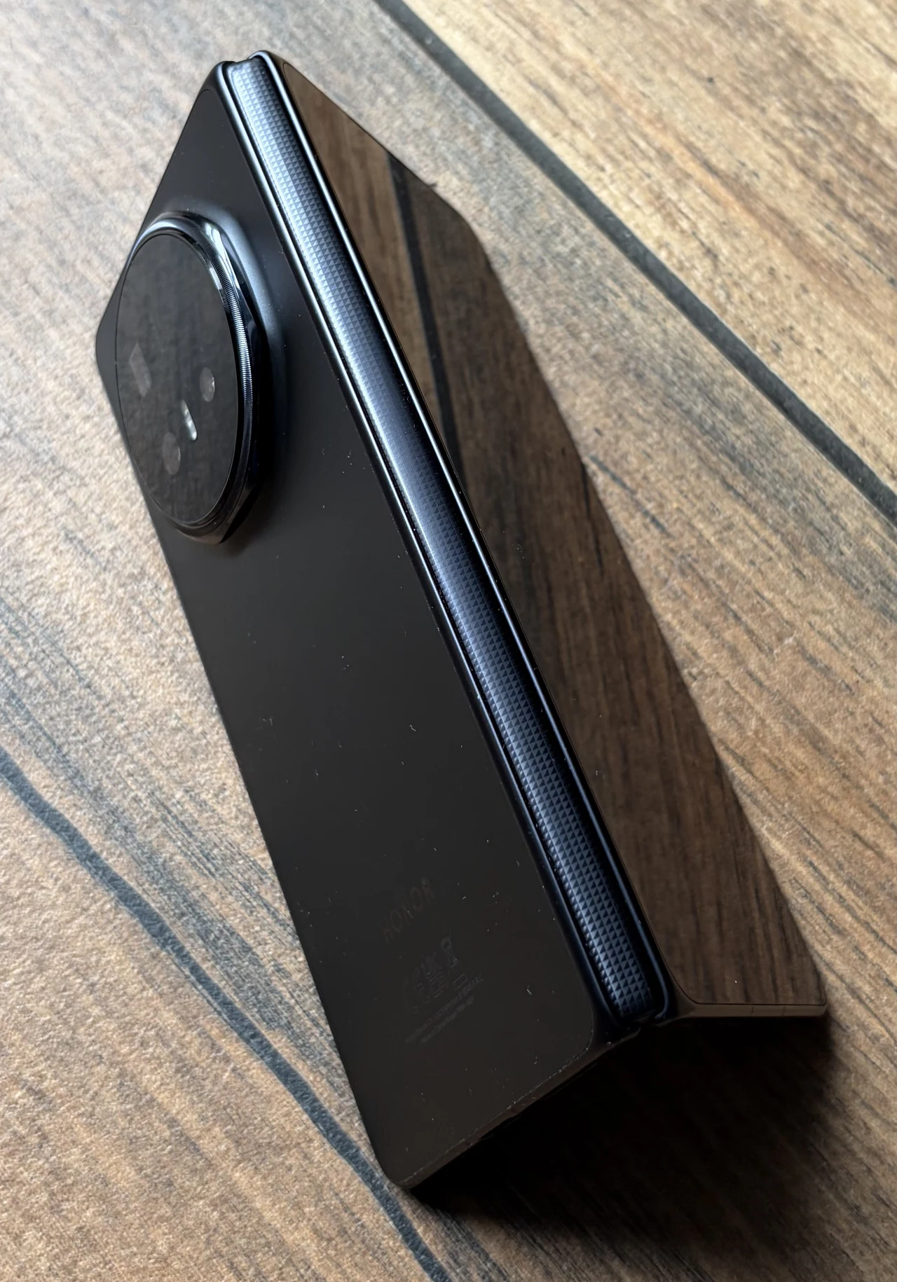 Składany smartfon w czarnej obudowie z dużą, okrągłą wyspą aparatów, leżący na powierzchni imitującej drewno.