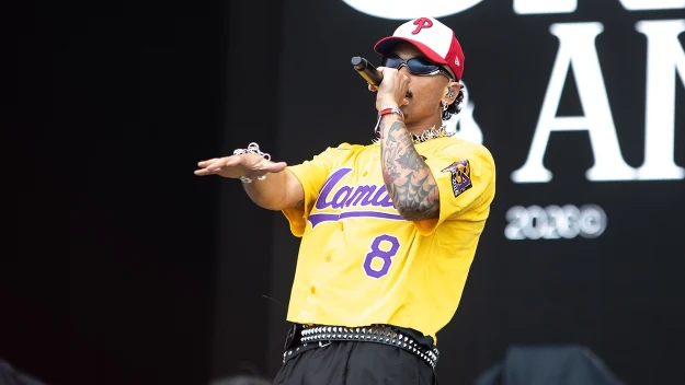 Mężczyzna w żółtej koszulce sportowej z numerem 8 i napisem „Lamar”, czerwonej czapce z daszkiem oraz okularach przeciwsłonecznych śpiewa na scenie, trzymając mikrofon i gestykulując ręką.