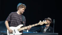 Mężczyzna grający na białej gitarze basowej na scenie, w tle muzyk siedzący przy klawiszach i noszący kapelusz.