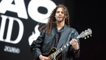 Mężczyzna z długimi, kręconymi włosami gra na czarnej gitarze elektrycznej, ubrany w czarną kurtkę skórzaną, na tle dużych liter tworzących element scenografii koncertowej.