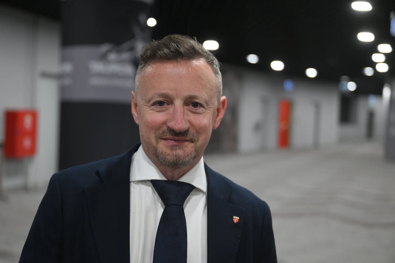 Mężczyzna w garniturze i granatowym krawacie stoi w nowoczesnym, przestronnym wnętrzu z jasnym oświetleniem sufitowym.