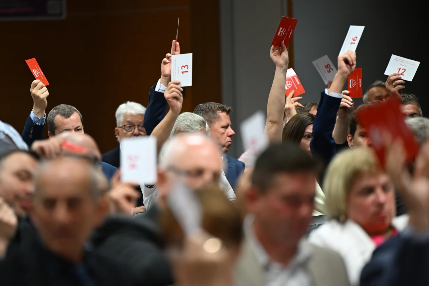 Grupa osób siedzących w sali, unoszących do góry czerwone kartki z numerami podczas głosowania.