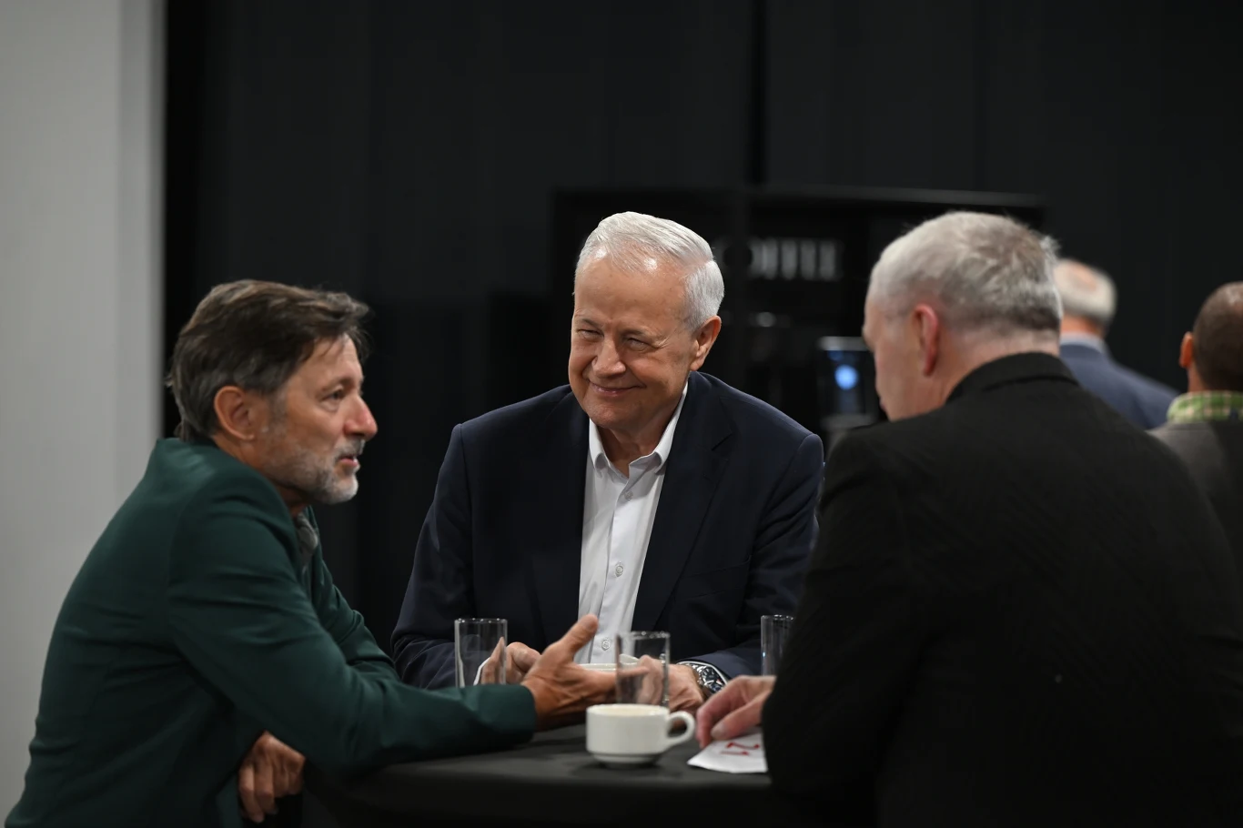 Trzech mężczyzn w garniturach siedzi przy okrągłym stole, rozmawiając ze sobą, przed nimi stoją szklanki z napojami i filiżanka kawy.