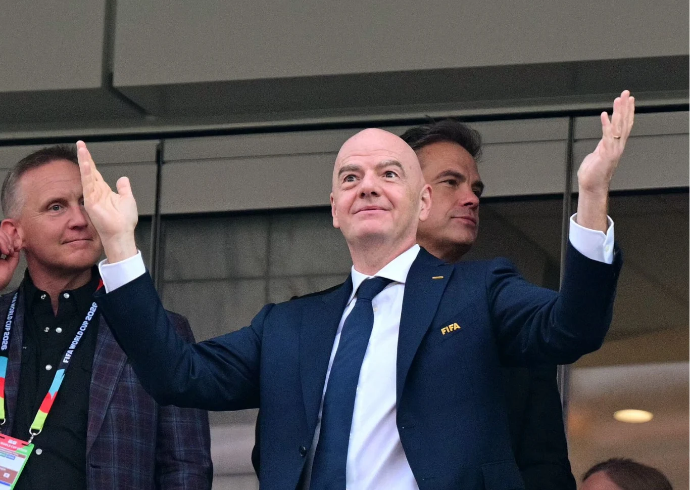 Mężczyzna w eleganckim garniturze z logotypem FIFA stoi z uniesionymi rękami, obok niego znajdują się inni mężczyźni przyglądający się wydarzeniu.