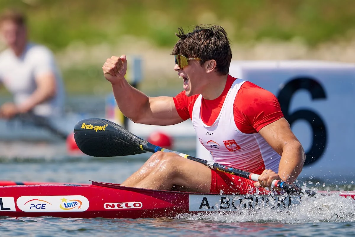 Kajakarz w biało-czerwonym stroju z uniesioną ręką świętuje sukces, siedząc w czerwonym kajaku na wodzie podczas zawodów sportowych.