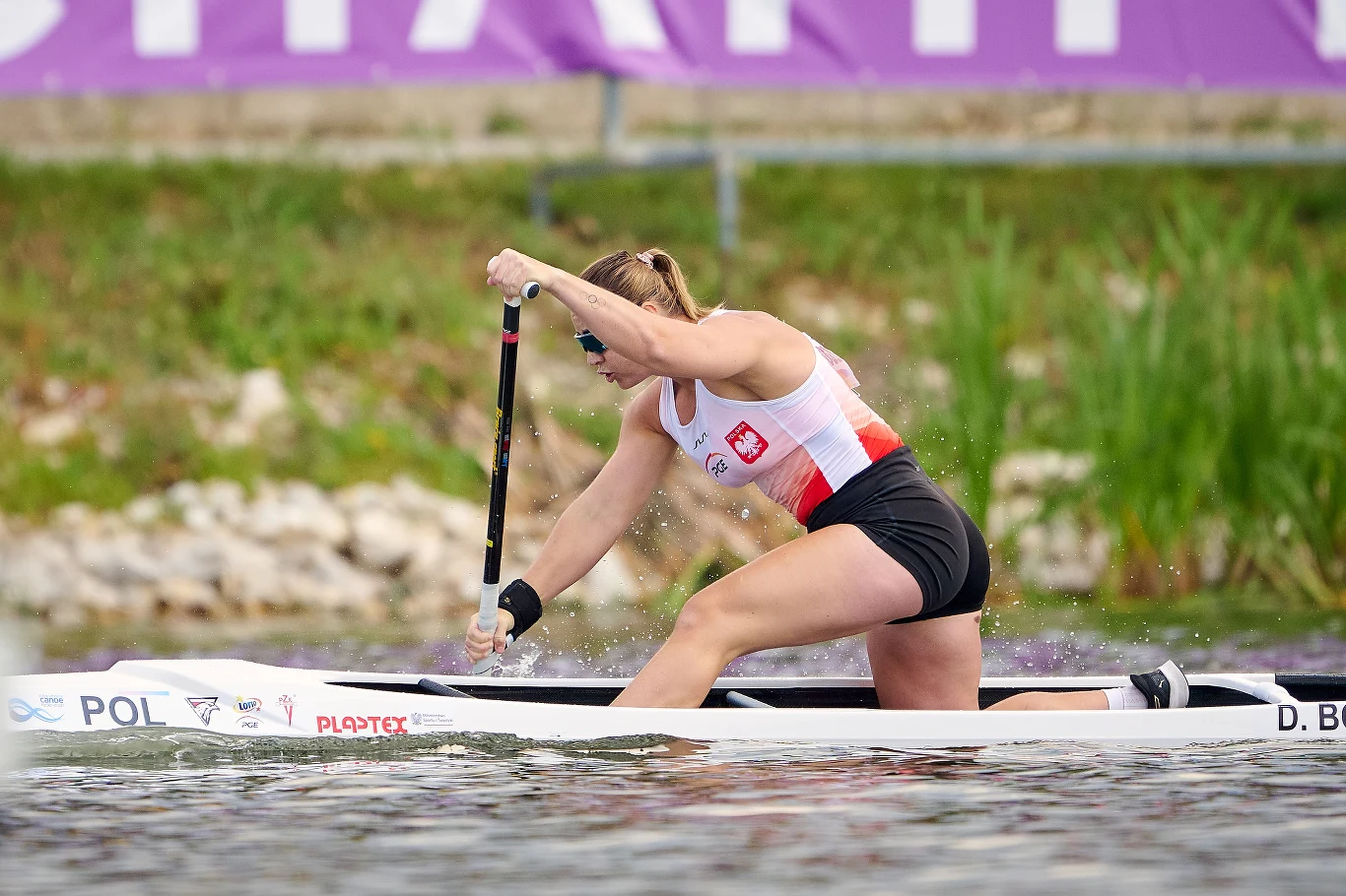 Zawodniczka w sportowej koszulce reprezentacji Polski energicznie wiosłuje na jednoosobowym kajaku, koncentrując się na wyścigu po spokojnej wodzie.