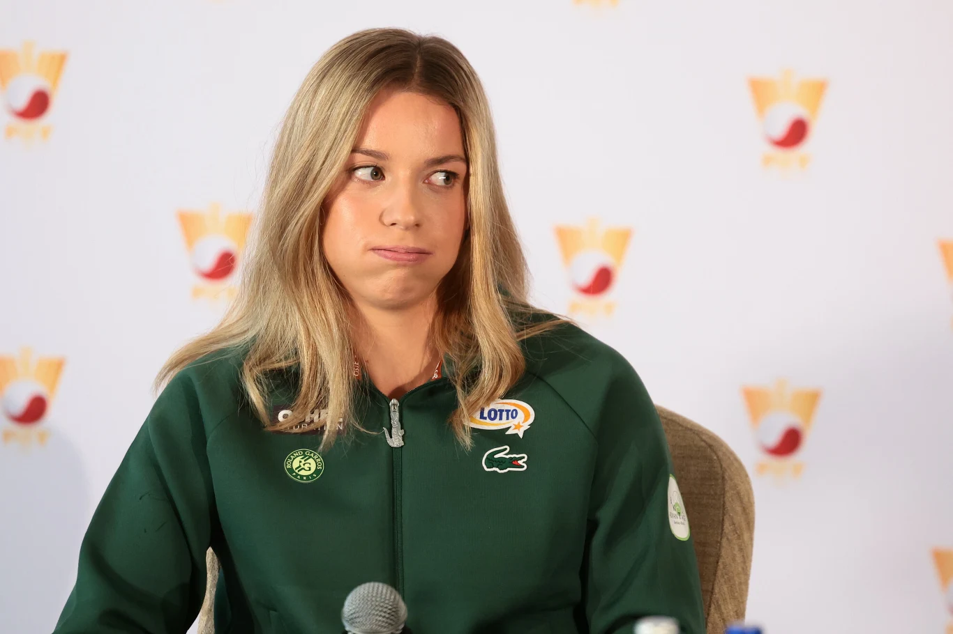Maja Chwalińska Kobieta w zielonej bluzie sportowej z logo sponsora, siedząca przy mikrofonie podczas konferencji prasowej, z wyrazem twarzy sugerującym niezadowolenie lub zakłopotanie.