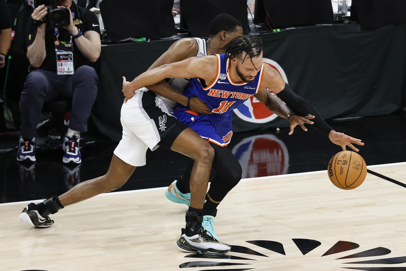 Kadr z meczu Knicks - Spurs Dwóch koszykarzy rywalizuje o piłkę podczas dynamicznej akcji na parkiecie, zawodnik w niebieskim stroju New York Knicks próbuje prowadzić piłkę, podczas gdy przeciwnik z drużyny San Antonio Spurs usiłuje odebrać mu piłkę, wykonując intensywną obronę.