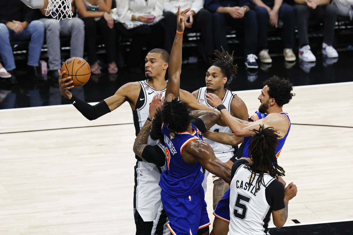 Kadr z meczu Knicks - Spurs Koszykarz w białym stroju unosi piłkę w kierunku kosza, jednocześnie próbując ominąć obrońcę w niebieskim stroju; obaj zawodnicy są otoczeni przez innych graczy, skupionych na walce pod koszem.