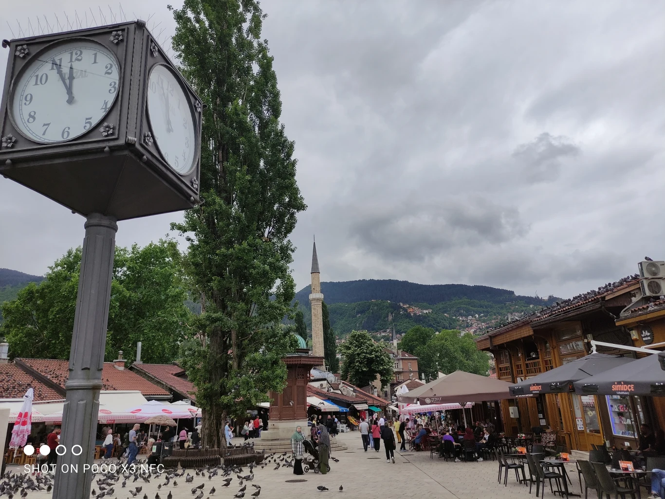 Zegar na słupie, w tle góry i minaret, po bokach liczne drewniane kawiarnie i parasole, na placu spacerujący ludzie oraz stado gołębi.