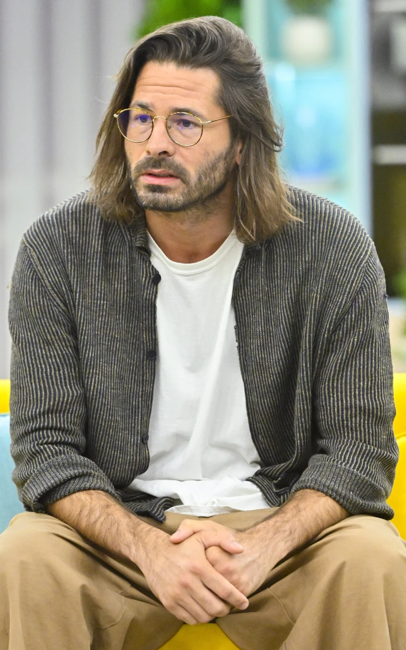 Mężczyzna z dłuższymi włosami i brodą, ubrany w jasny t-shirt i rozpinany sweter, siedzi na żółtej sofie z rękami splecionymi na kolanach, nosi okulary o okrągłych oprawkach.
