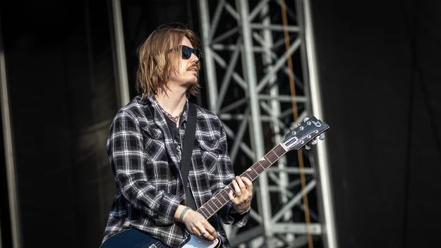 Mężczyzna w kraciastej koszuli i okularach przeciwsłonecznych gra na elektrycznej gitarze podczas koncertu na otwartej scenie.