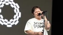 Wokalista śpiewający do mikrofonu na scenie, ubrany w jasny t-shirt z czarnym nadrukiem, w tle abstrakcyjny biały wzór na ciemnym ekranie.