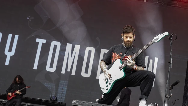 Gitarzysta w czarnej koszulce i białych butach gra na gitarze basowej na scenie podczas koncertu rockowego, z tyłu widoczny drugi muzyk z gitarą elektryczną na tle dużego ekranu z fragmentem napisu.