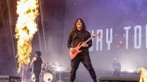 Gitarzysta z długimi włosami gra na pomarańczowej gitarze elektrycznej na dużej scenie, obok wybuchających płomieni, podczas koncertu zespołu rockowego; w tle bębniarz i inni muzycy.