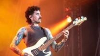 Mężczyzna z tatuażami gra na gitarze basowej na scenie koncertowej, w tle intensywne światło w odcieniach pomarańczu i żółci.