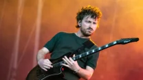 Mężczyzna z brodą i kręconymi włosami grający na elektrycznej gitarze z wyrazem skupienia na twarzy, oświetlony kolorowym światłem scenicznym.