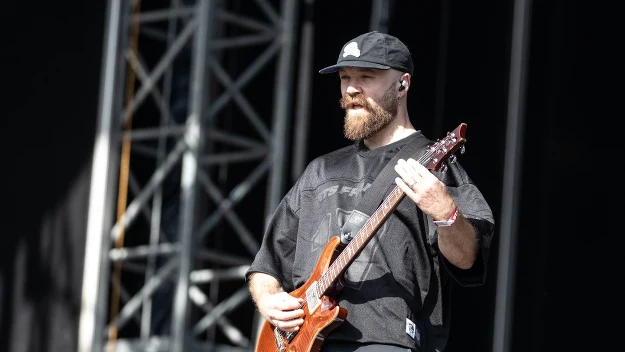 Mężczyzna z brodą w czarnej czapce i oversize'owej koszulce gra na brązowej gitarze elektrycznej na scenie koncertowej.