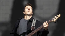 Gitarzysta basowy w ciemnej kurtce gra na scenie, trzymając instrument i patrząc w górę z poważnym wyrazem twarzy.