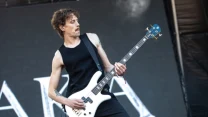 Mężczyzna w czarnym podkoszulku grający na białej gitarze basowej podczas występu scenicznego, na tle dużej grafiki z widocznymi literami.