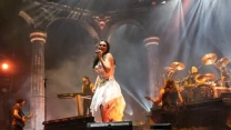 Wokalistka w białej sukni śpiewająca na scenie podczas koncertu rockowego, otoczona przez muzyków grających na gitarach i perkusji, w tle podświetlane kolumny scenografii.