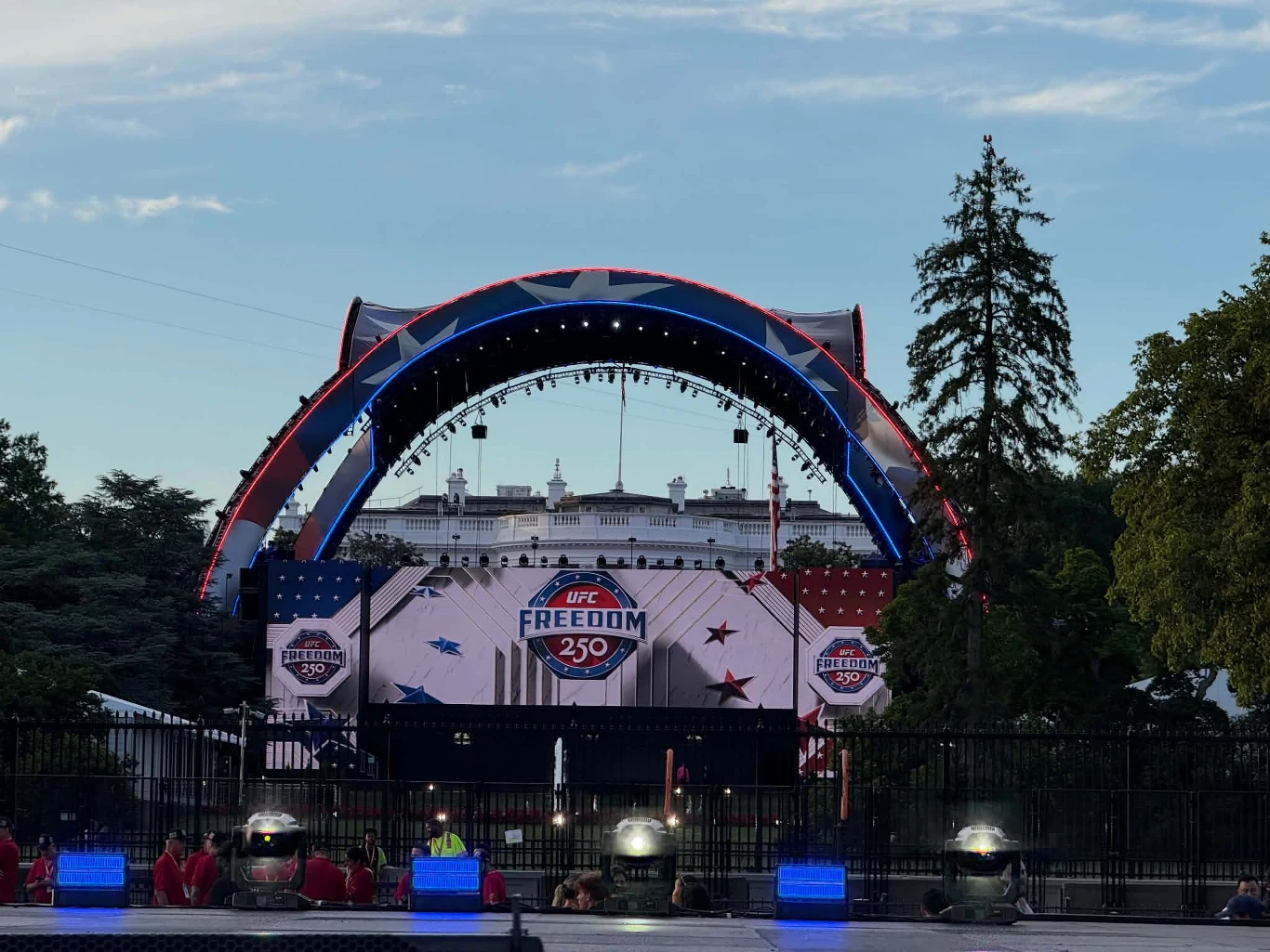 Duża scena z łukowatą konstrukcją i ekranem LED z napisem „UFC Freedom 250” ustawiona na tle białego, imponującego budynku przypominającego rezydencję rządową, otoczona drzewami oraz zabezpieczeniami barierek i ochroną po bokach.