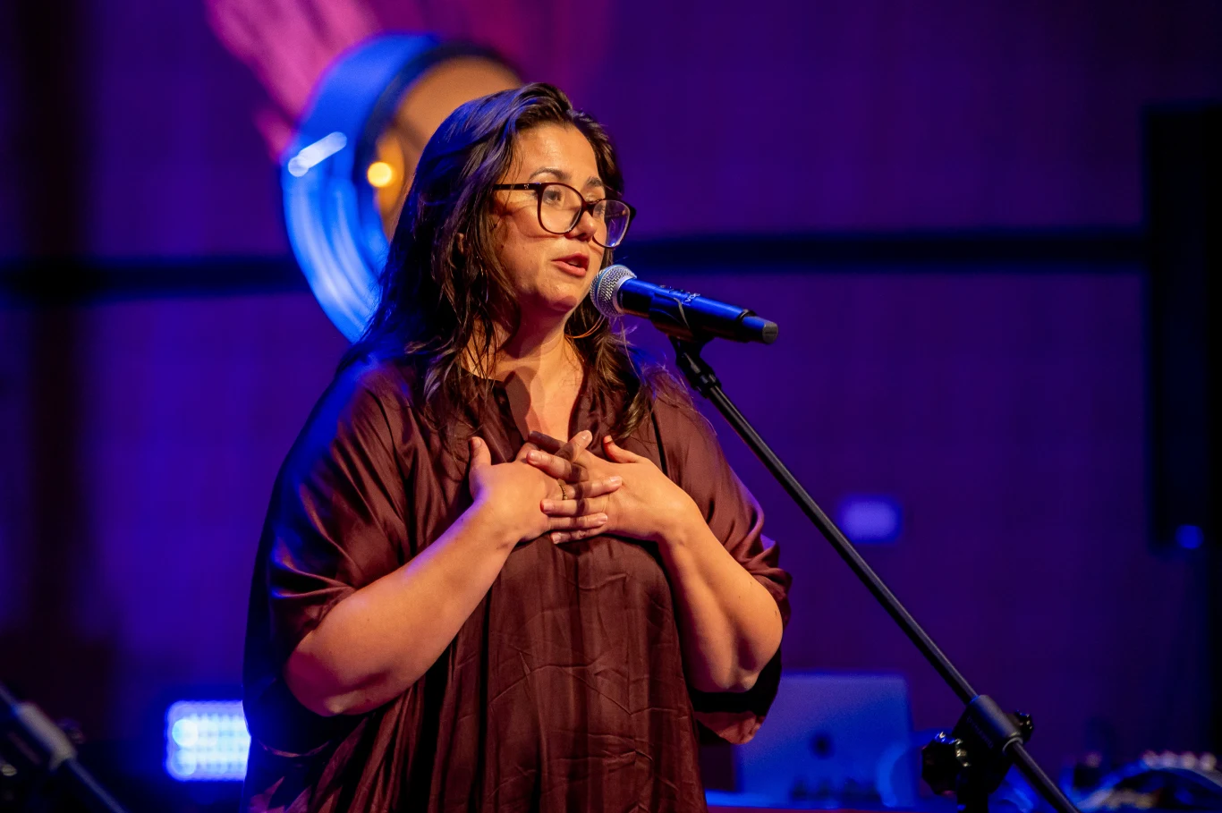 Anna Serdiukow, dyrektorka programowa festiwalu "Młodzi i Film" Kobieta z długimi włosami i okularami śpiewa lub przemawia do mikrofonu, trzymając dłonie na klatce piersiowej, oświetlona kolorowym światłem sceny.