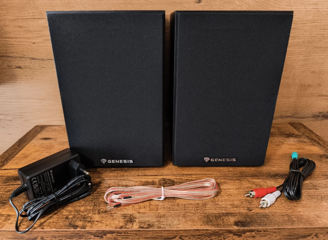 Dwie czarne kolumny głośnikowe Genesis ustawione obok siebie, przed nimi zasilacz, przewód głośnikowy oraz komplet kabli audio zakończonych wtykami RCA i mini jack.