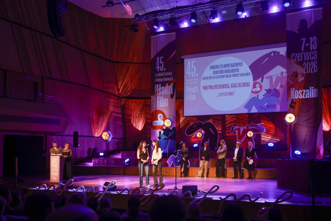 "Młodzi i Film" 2026: Martyna Byczkowska i Kaja Zalewska z nagrodą za film "La petite mort" Uroczysta gala filmowa na dużej scenie, kilka osób wręcza nagrody, a w tle wyświetlony jest duży ekran z informacjami o wydarzeniu oraz dekoracje świetlne i banery festiwalowe.